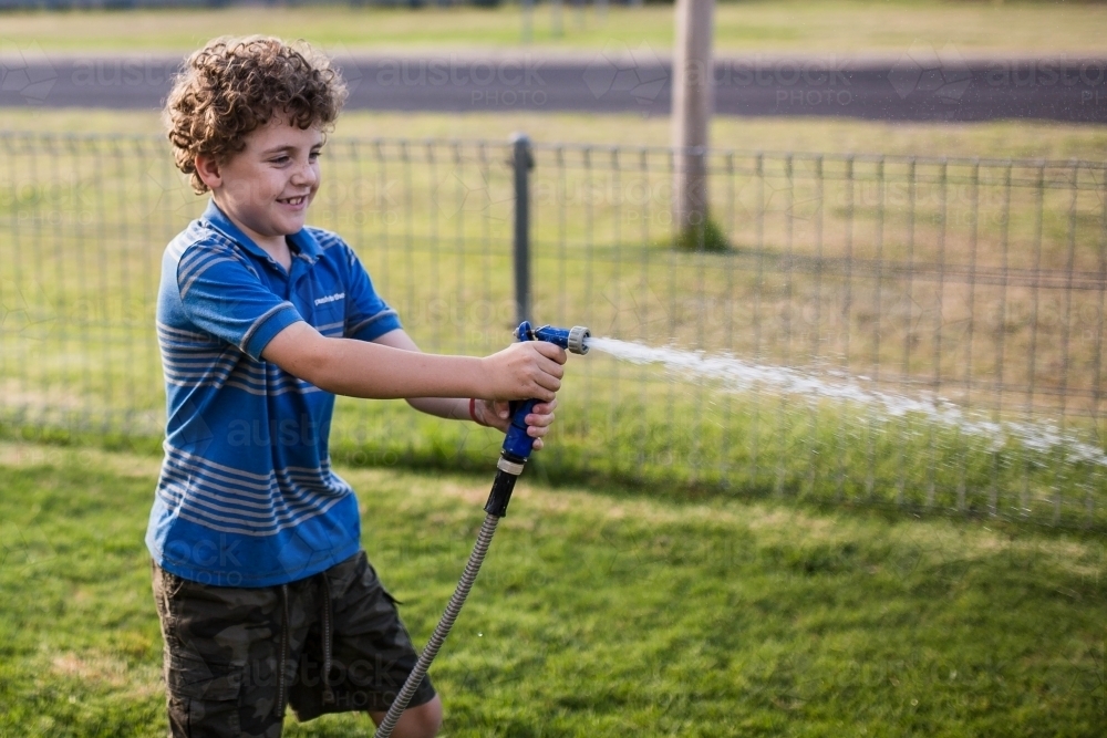 Boy smiling pointing water hose on lawn - Australian Stock Image