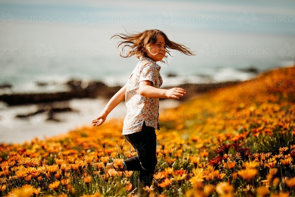 Boy spinning in field of flowers on clifftop next to the ocean - Australian Stock Image