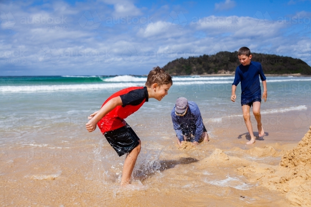 Boy splashing in water at the beach beside sand castle - Australian Stock Image