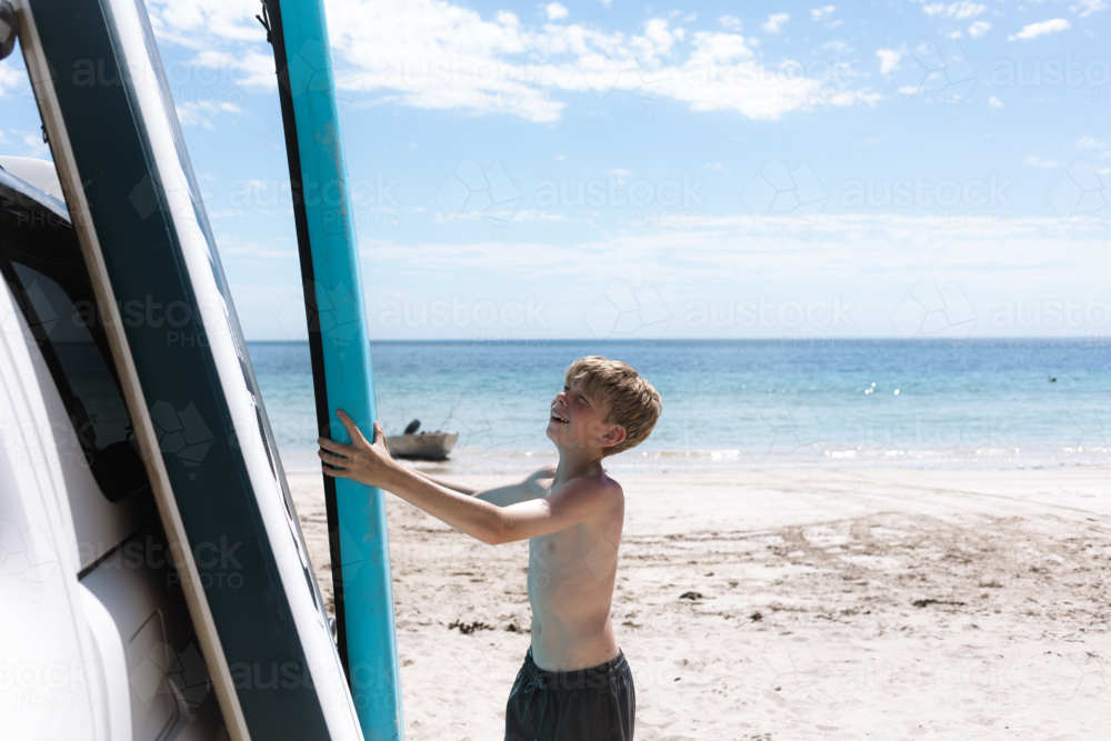 Boy stacking paddle boards against car on clear blue beach in full summer sun - Australian Stock Image