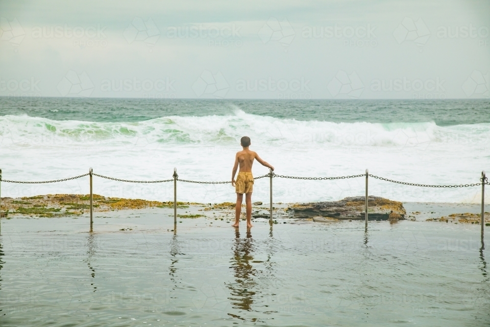 Boy standing at the fence on the edge of the Bogey Hole in Newcastle, NSW on an overcast day - Australian Stock Image
