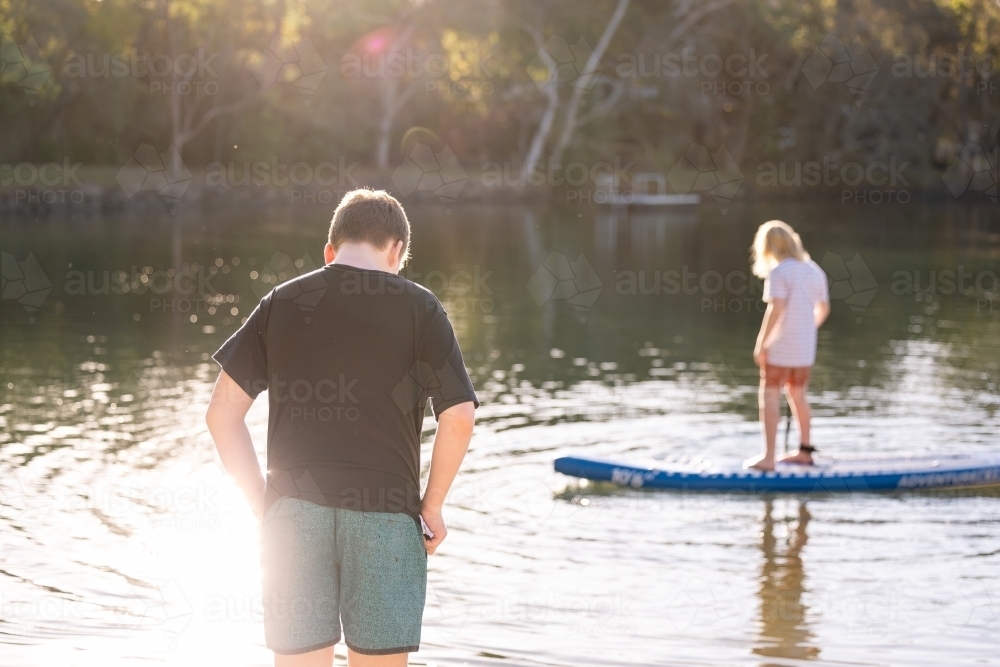 Boy standing in golden afternoon sun with brother on SUP in background - Australian Stock Image