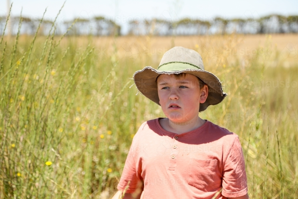 Boy standing in long grass on farm wearing green hat on hot summer day - Australian Stock Image