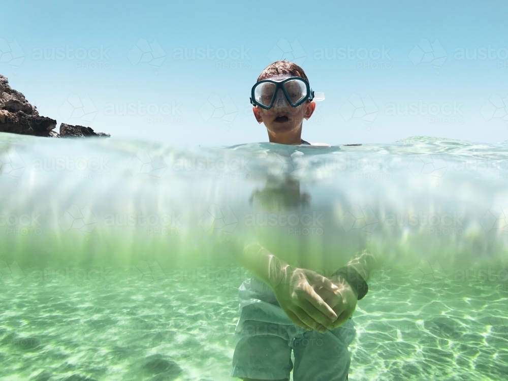 Boy standing in ocean with view underwater of body and above water of head wearing snorkel mask - Australian Stock Image