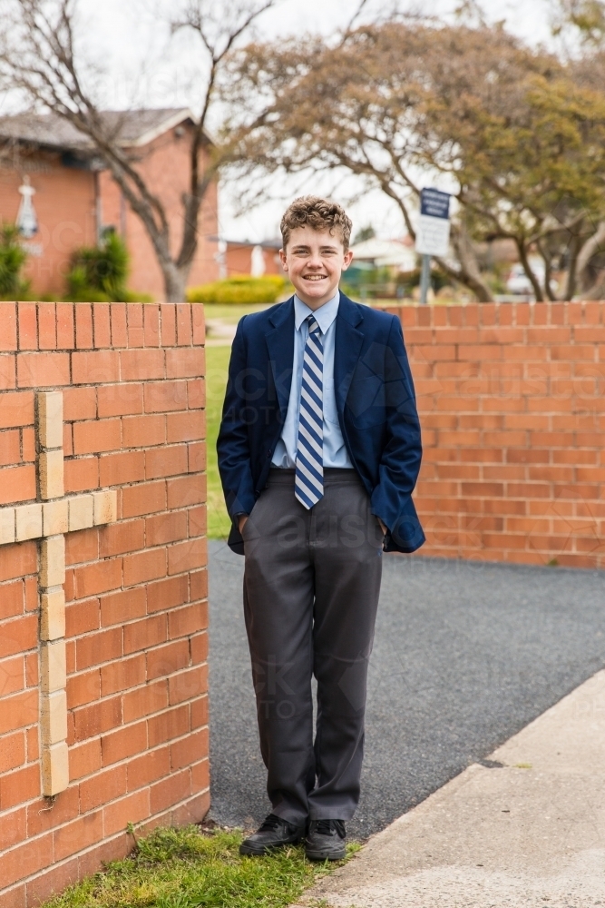 Boy standing in school uniform near brick entrance with cross smiling - Australian Stock Image