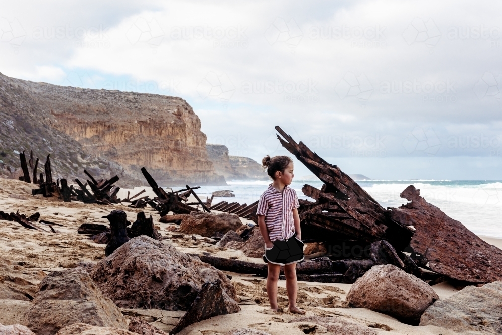 Boy standing on beach next to shipwreck with ocean and cliffs behind - Australian Stock Image