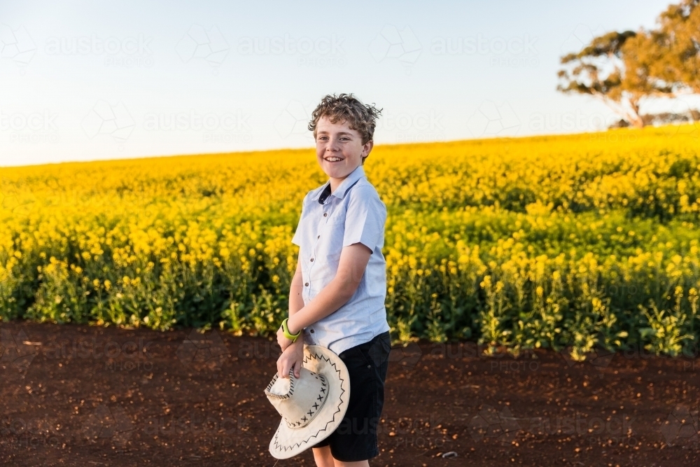 Boy standing on dirt road holding hat smiling near canola field on farm - Australian Stock Image