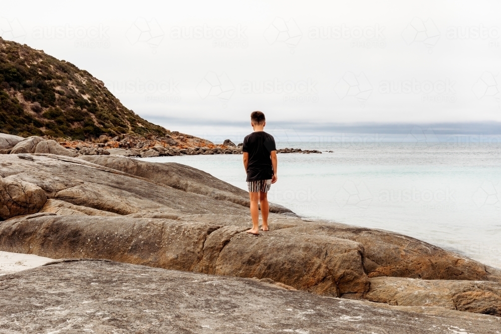 Boy standing on rocks looking at the ocean - Australian Stock Image