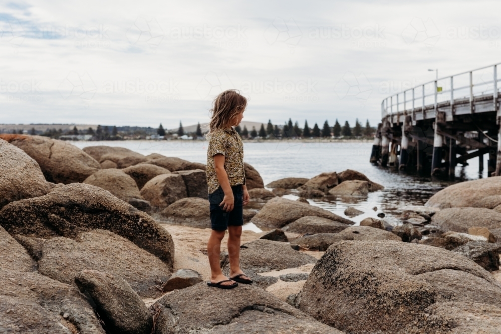 Boy standing on rocks looking at the ocean next to a jetty - Australian Stock Image