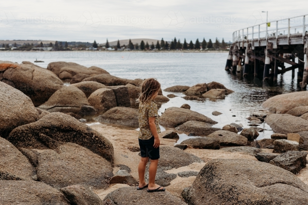 Boy standing on rocks next to the ocean and jetty - Australian Stock Image
