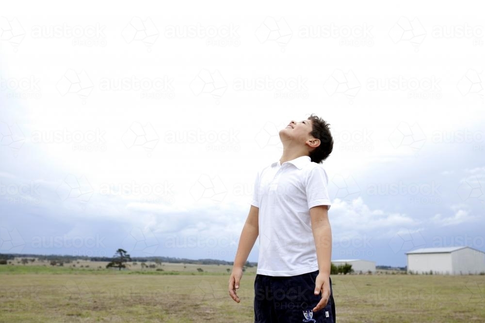 Boy standing outside in a farm paddock looking up at sky - Australian Stock Image