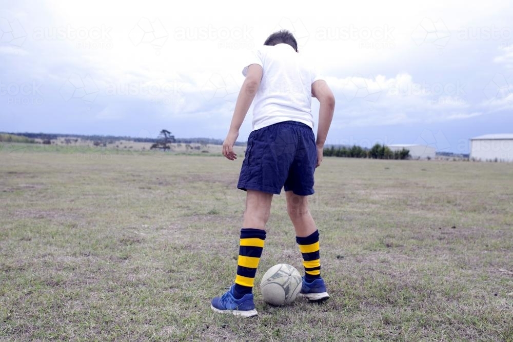 Boy standing over a football in a rural paddock - Australian Stock Image