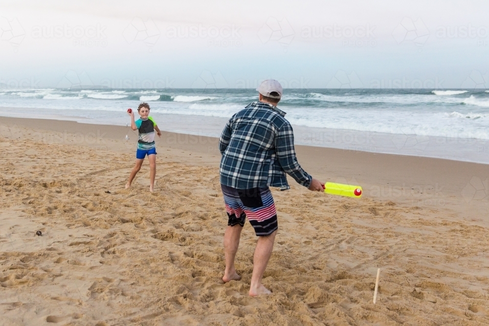 Boy throwing ball to his father holding bat playing cricket on the beach - Australian Stock Image