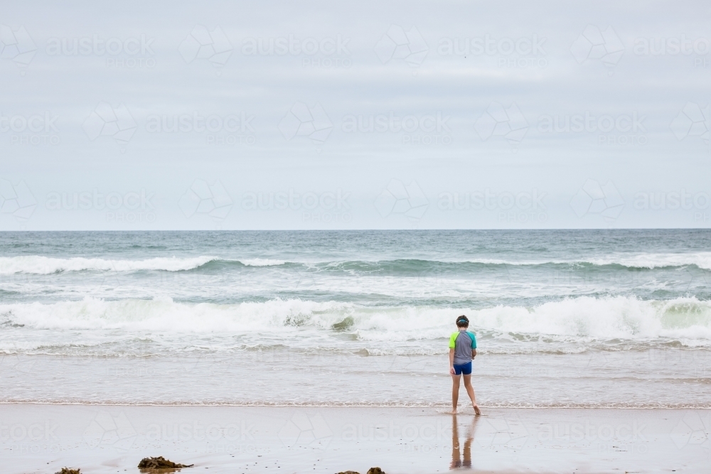 Boy walking alone on beach towards waves - Australian Stock Image