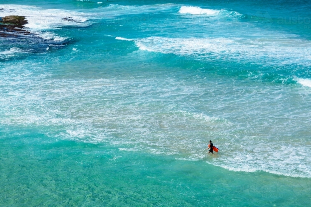 Boy walking in the water holding a body board at the beach - Australian Stock Image
