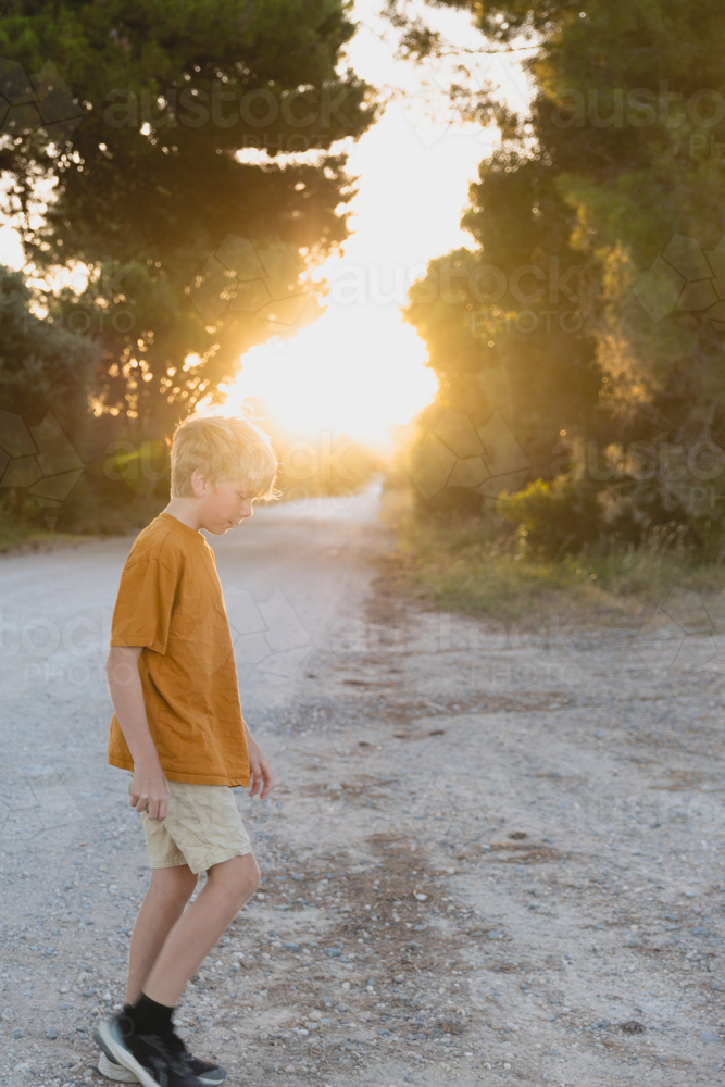 Boy walking on dirt country road at sunset wearing orange top and neutral shorts - Australian Stock Image