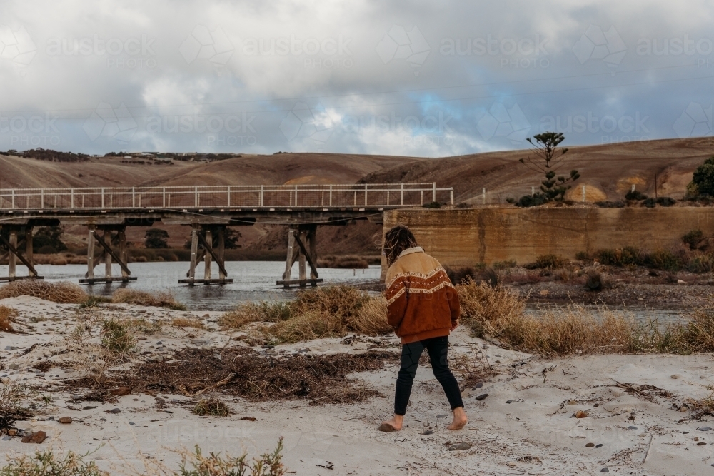 Boy walking on the beach with a bridge and hills in the distance - Australian Stock Image