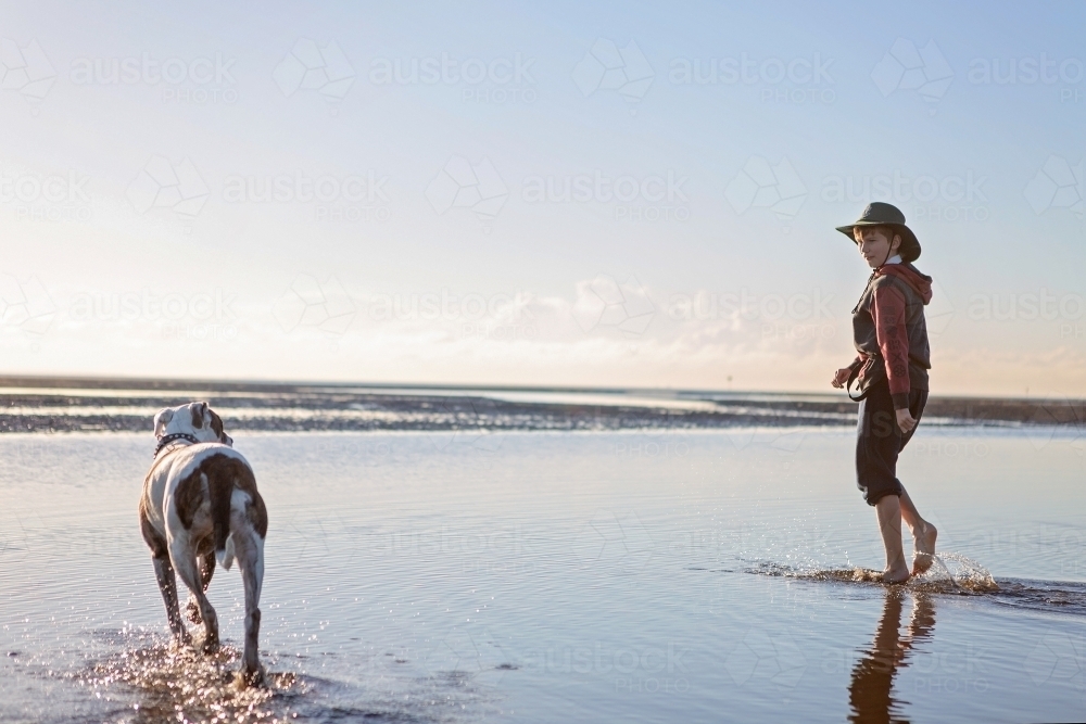 Boy walks on the beach at lowtide in winter barefoot with his dog - Australian Stock Image