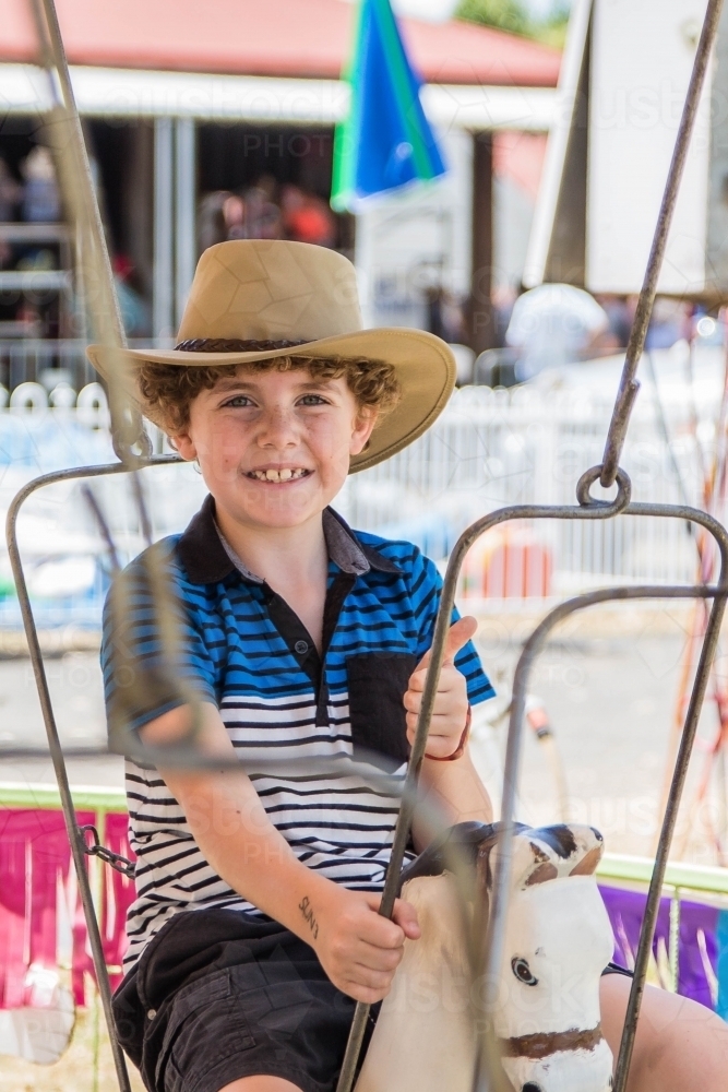 Boy wearing akubra hat sitting on horse ride at local show with thumbs up smiling - Australian Stock Image