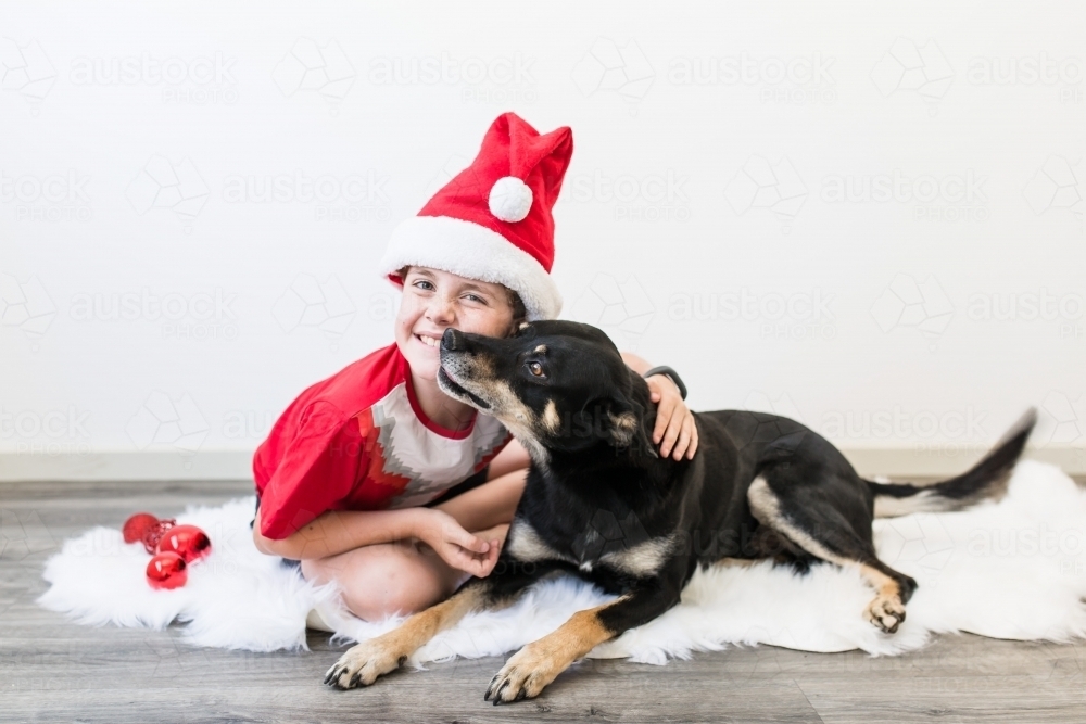 Boy wearing santa hat sitting on rug being kissed by kelpie dog at Christmas - Australian Stock Image