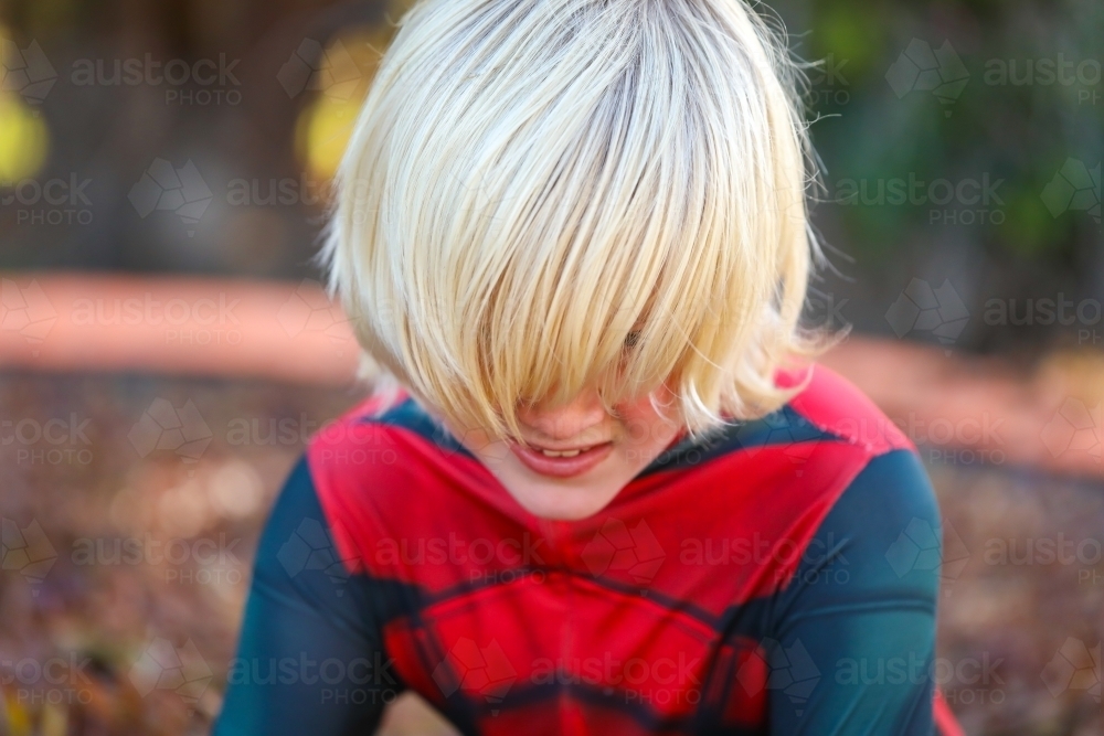 Boy wearing superhero suit with long blonde hair covering eyes - Australian Stock Image
