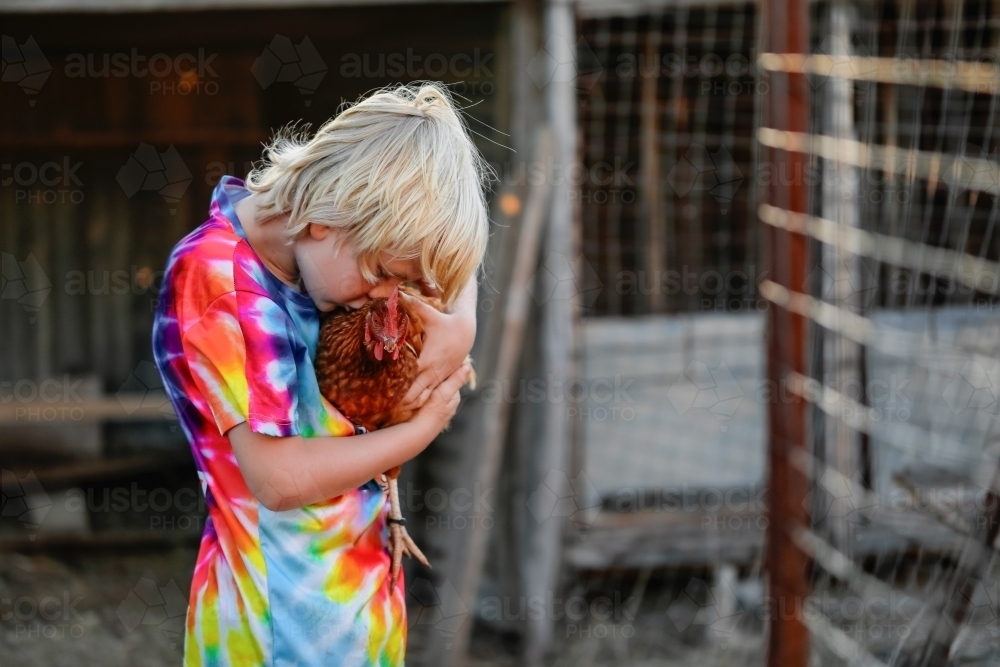 Boy wearing vibrant rainbow shirt holding Isa Brown chicken on farm - Australian Stock Image