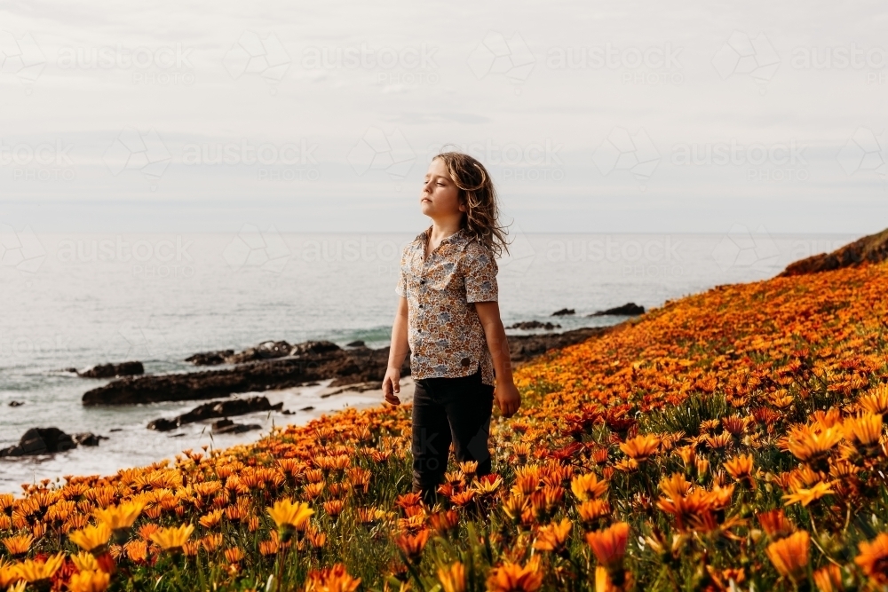 Boy with eyes closed and face to the sun standing in a field of flowers on a clifftop next to ocean - Australian Stock Image