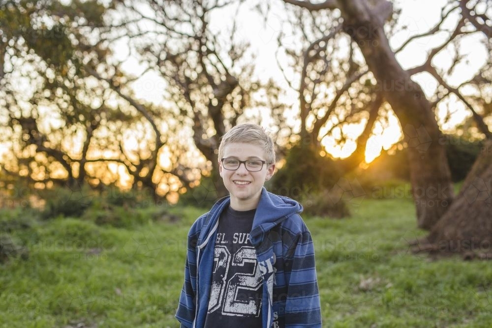 Boy with glasses standing in front of trees in afternoon sun - Australian Stock Image