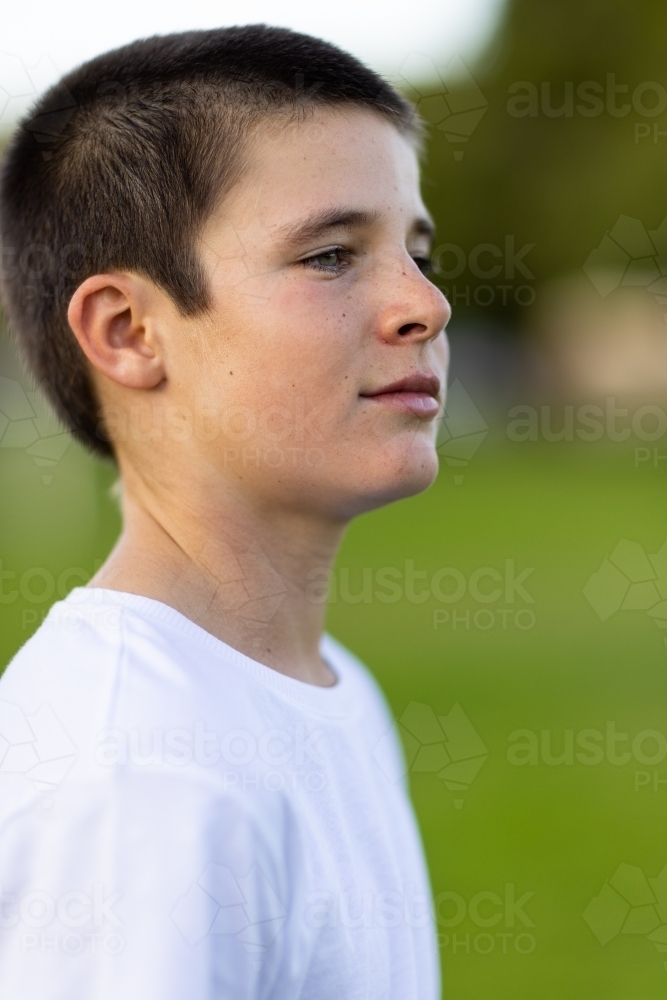 boy with short hair wearing white tee shirt against green outdoor blurry background - Australian Stock Image