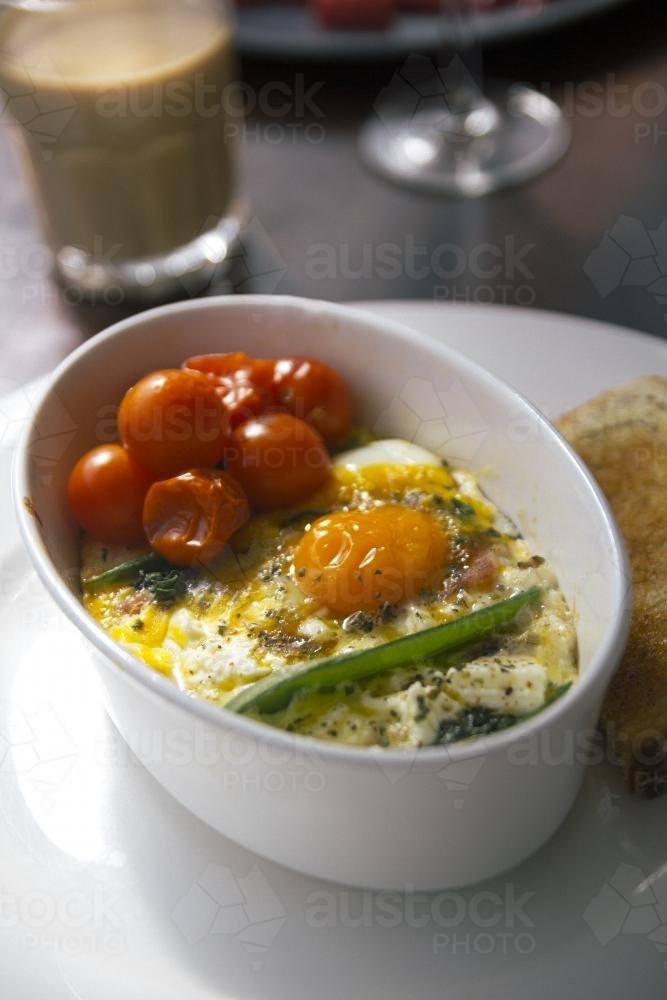 Breakfast of baked eggs and cherry tomatoes in a white bowl at a cafe - Australian Stock Image