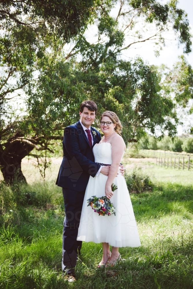 Bride and Groom standing beside a tree in paddock looking at camera - Australian Stock Image