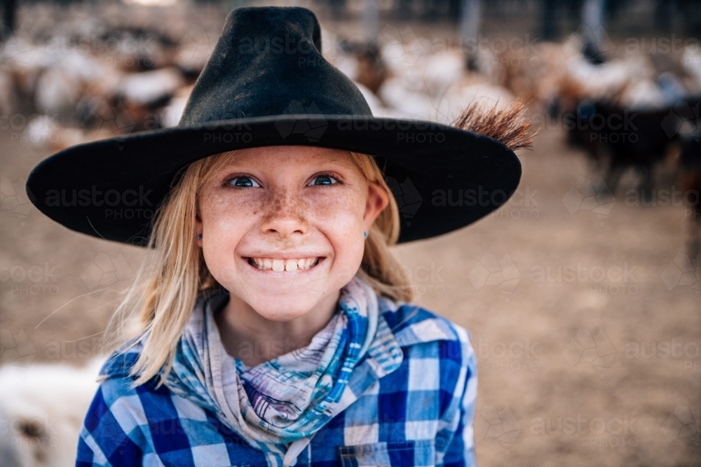 Bright eyed smiling girl with hat, checkered shirt and scarf with goats in the background - Australian Stock Image
