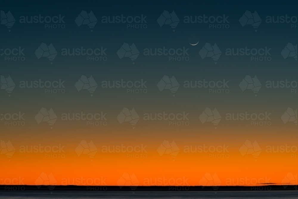 Bright orange and dark blue post sunset sky with crescent moon and low horizon - Australian Stock Image