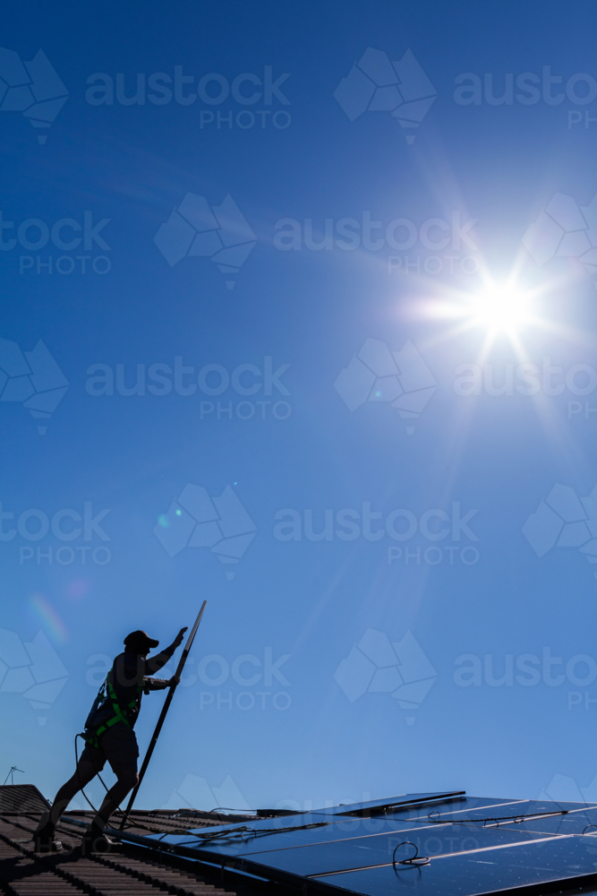 Bright sun flare in blue sky over person installing solar panels on roof of home - Australian Stock Image