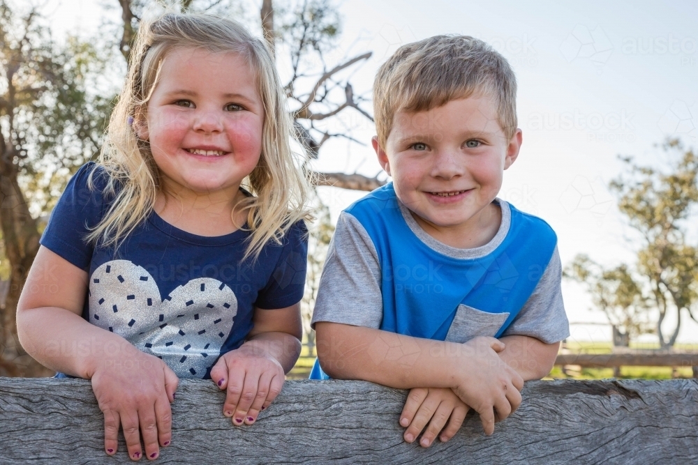 Brother and sister leaning on wooden fence smiling - Australian Stock Image