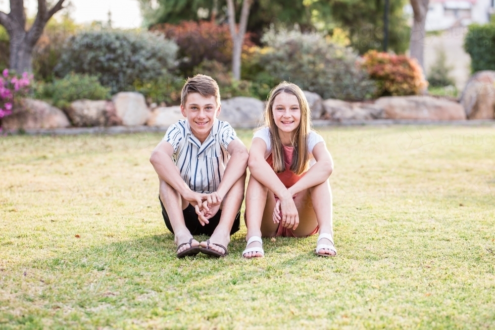 Brother and sister sitting close together resting arms on knees smiling - Australian Stock Image