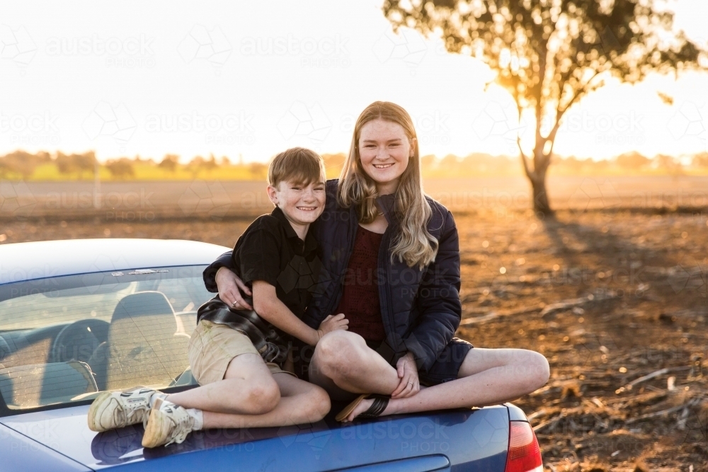 Brother and sister sitting on boot of car at sunset in dirt paddock on farm - Australian Stock Image