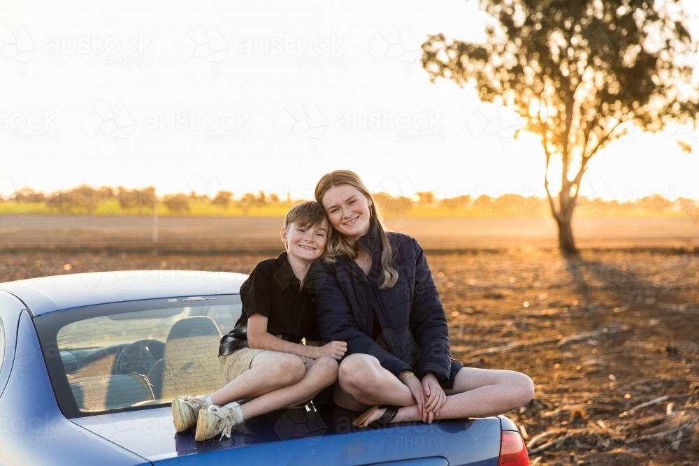 Brother and sister sitting on boot of car smiling in dirt paddock on farm at sunset - Australian Stock Image