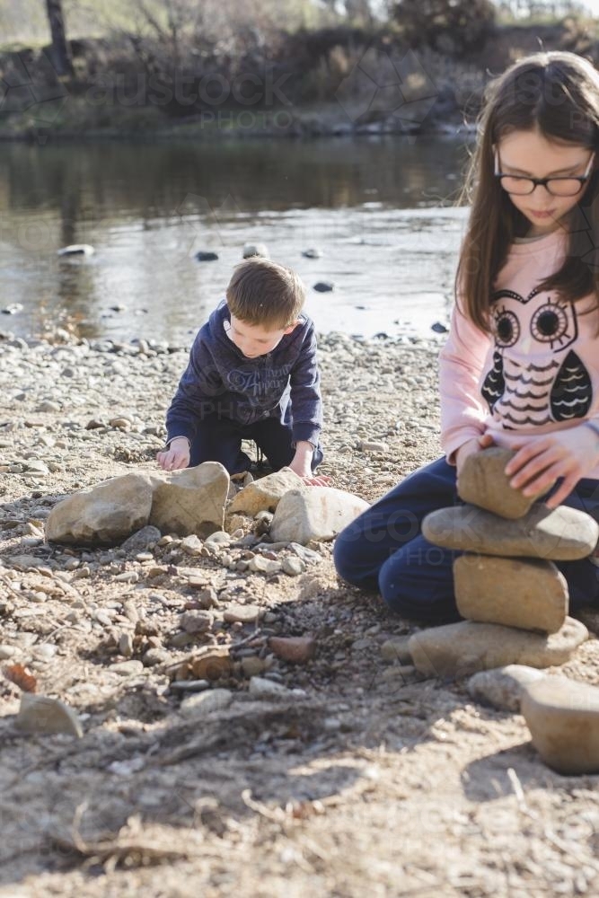Brother and sister stacking rocks by rivers edge - Australian Stock Image