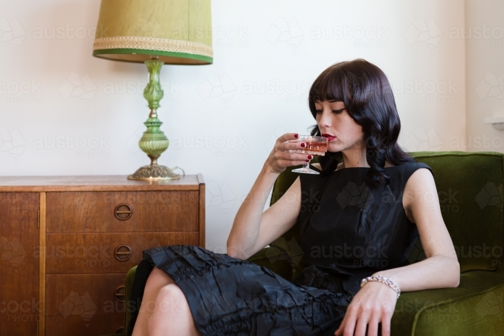 Brunette woman sipping pink champagne in a retro living room - Australian Stock Image