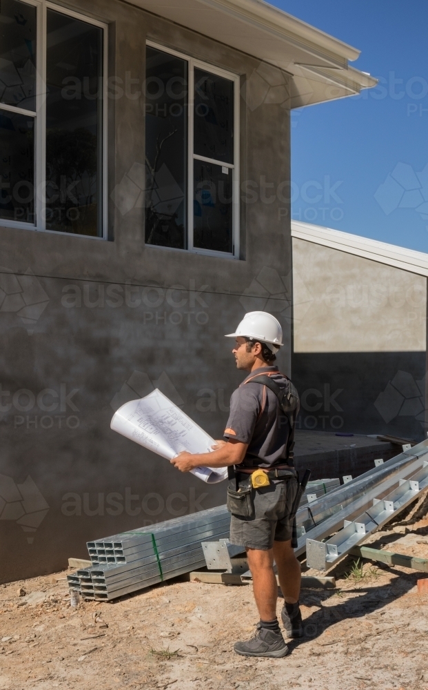 Builder with hard hat & tool belt holding plans outside new house construction - Australian Stock Image