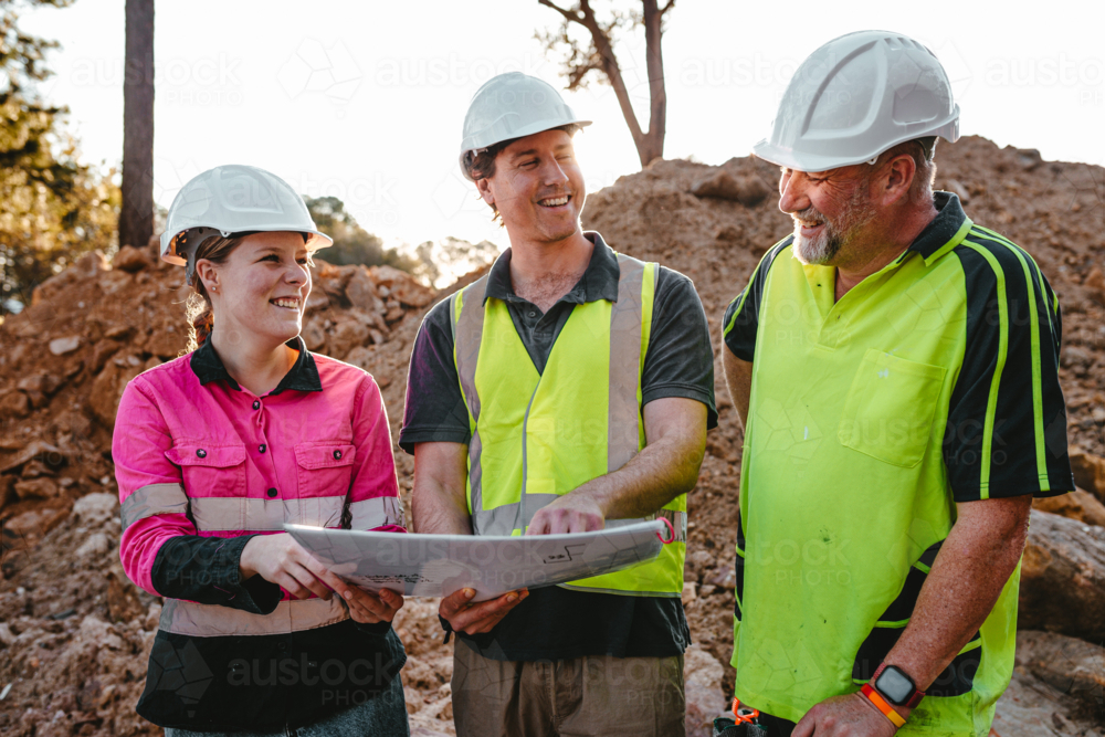 Builders in hardhats smiling and discussing job while holding a blueprint together on worksite - Australian Stock Image