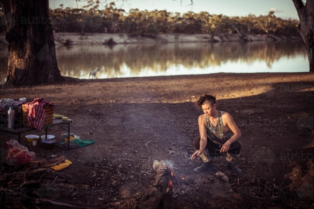Camping life - person squatted by a campfire next to a river - Australian Stock Image
