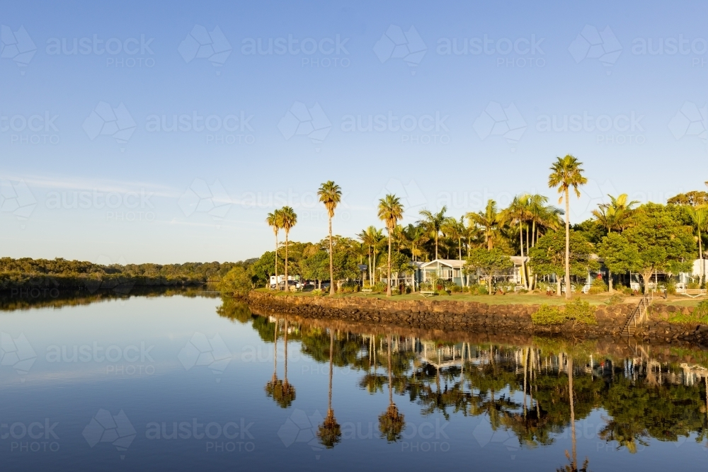 Caravan park beside Brunswick River with tall palm trees reflected in water - Australian Stock Image