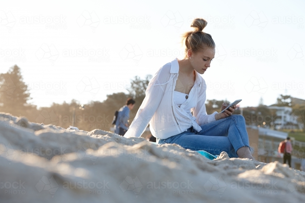 Carefree young blonde-haired woman at beach checking mobile phone - Australian Stock Image