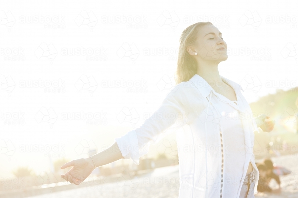 Carefree young blonde-haired woman at beach with arms out-stretched enjoying the sunshine - Australian Stock Image