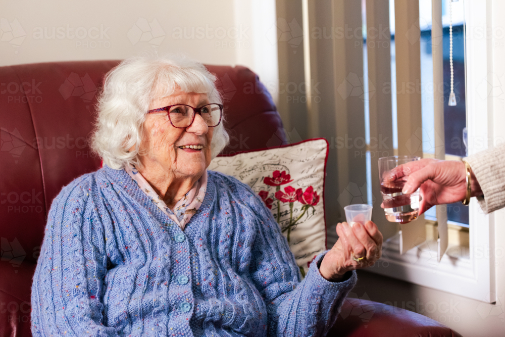 Carer passing glass of water in cup to elderly Australian woman holding medication tablets - Australian Stock Image