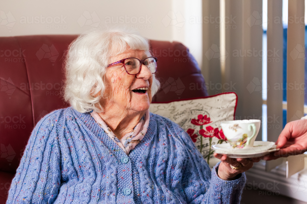 Carer passing tea in mug to elderly Australian woman sitting on armchair smiling - Australian Stock Image