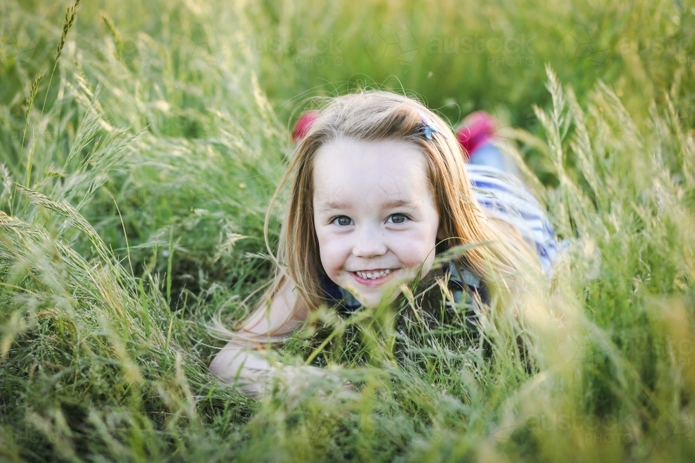 Caucasian girl with long hair lying in long grass at the park with a big smile - Australian Stock Image