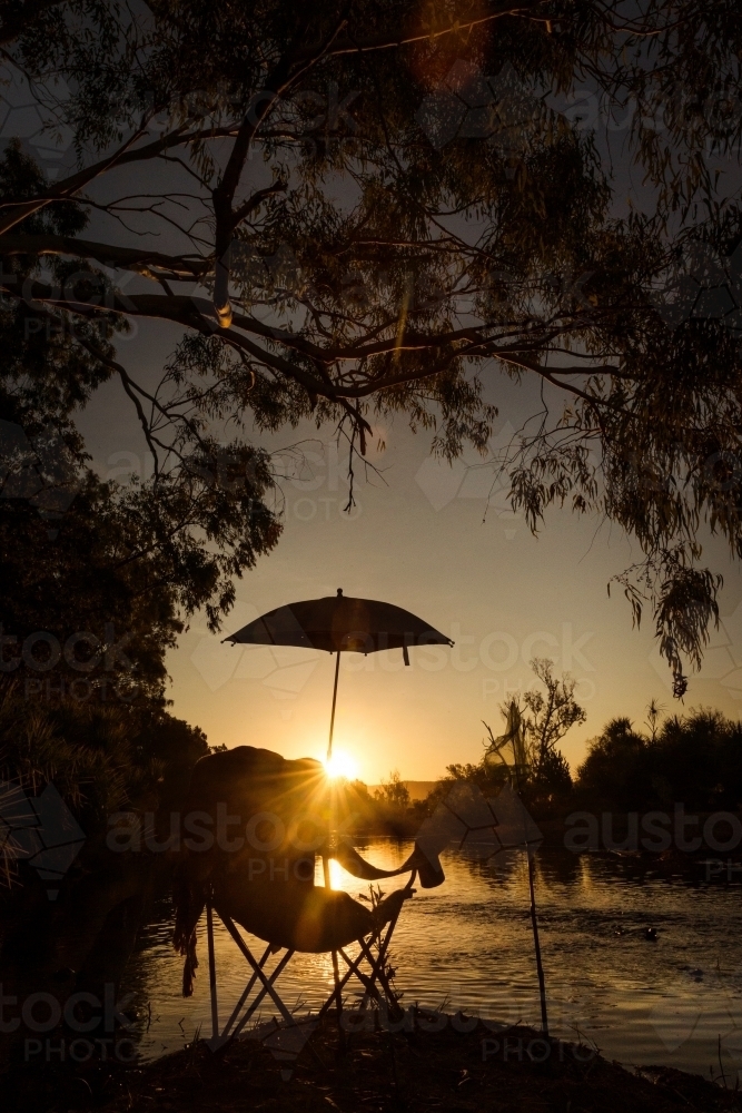 Chair with umbrella during sunrise with orange sky in front of creek with trees - Australian Stock Image
